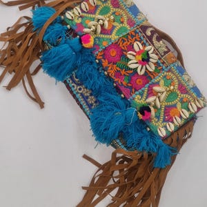 May include: A vibrant patchwork clutch bag with detailed embroidery, seashells, and blue tassels. The bag has brown suede fringe and a long strap. The design incorporates bright floral patterns and a variety of colours, including pink, green, and blue.