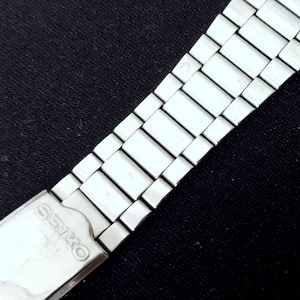 May include: A silver-toned stainless steel watch bracelet featuring a series of rectangular links and a folding clasp. The metal clasp is engraved with the brand name SEIKO in capital letters. The watch band has a polished finish.