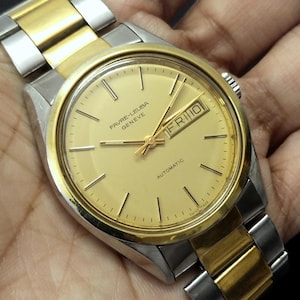 May include: A gold and silver Favre-Leuba Geneve automatic wristwatch. The watch face is gold with black markers and hands. The day and date are displayed at the 3 o'clock position. The watch band is a combination of gold and silver links.