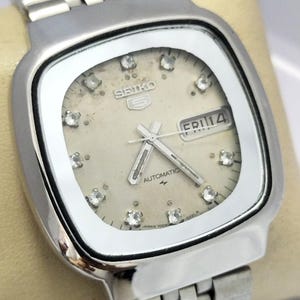 Puede incluir: Un reloj de pulsera automático Seiko 5 plateado con caja cuadrada y brazalete de eslabones metálicos. La esfera color crema presenta marcadores de hora de cristal, manecillas plateadas y una ventana de día y fecha con el texto FRI 14.