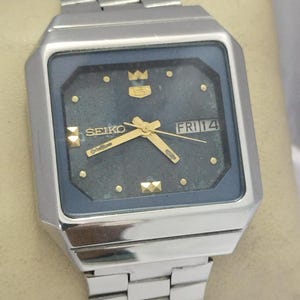 Può includere: Un orologio da polso Seiko color argento con cassa quadrata e quadrante scuro testurizzato. L'orologio presenta lancette dorate, indici dorati e una finestrella giorno-data che mostra FRI 14. Include un cinturino a maglie in metallo abbinato.