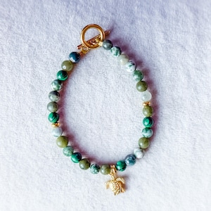 May include: A bracelet made of green and white beads, with gold-coloured accents and a turtle charm. The bracelet has a gold-coloured toggle clasp. The beads are displayed on a white surface.
