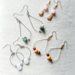 May include: An assortment of handmade earrings featuring various designs. Some earrings have gold or silver hooks, with teardrop, hoop, and linear shapes. The earrings are adorned with beads in shades of pink, white, green, and brown, and some have seashell charms.