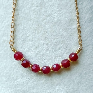 May include: A gold chain necklace featuring faceted, deep red beads. The necklace has a delicate gold chain and is displayed against a soft, light background. The beads are evenly spaced and add a pop of color.