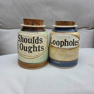 May include: Two ceramic jars with cork stoppers. The left jar is light green and brown with the text Shoulds & Oughts in a dark oval. The right jar is cream and blue with the text Loopholes in a dark oval. Both feature a speckled finish.