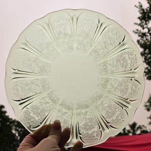 May include: A round, pale yellow translucent glass plate featuring a scalloped edge and a decorative embossed floral pattern. The design consists of radiating panels separated by smooth, raised ribs that extend from the center to the rim.