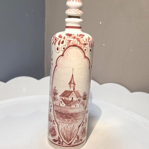 Può includere: Una bottiglia cilindrica in ceramica con tappo a punta abbinato, caratterizzata da una base bianca con motivi floreali dipinti a mano in marrone rossiccio e un'illustrazione centrale incorniciata di una chiesa vicino all'acqua.
