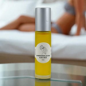 May include: A roll-on bottle with a silver lid and a yellow liquid. The label says "Aphrodisiac Elixir Gaiae" with a botanical design. The bottle sits on a glass surface, with a blurred background.