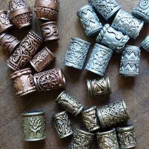 May include: A collection of silver, copper, and bronze hair beads with intricate designs. The beads are round and cylindrical, with some featuring a tree of life motif.