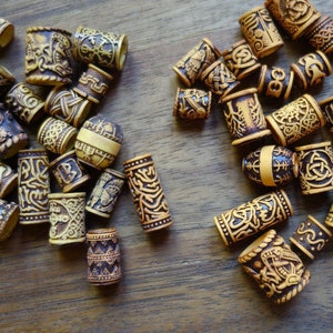 May include: A collection of wooden hair beads with intricate carved designs. The beads are in shades of brown and beige, and some have a darker brown finish. The beads are scattered on a wooden surface.