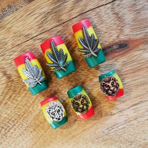 Puede incluir: Seis cuentas cilíndricas de colores con diferentes diseños metálicos. Las cuentas son rojas, amarillas y verdes, y presentan diseños de hojas de marihuana y cabeza de león.