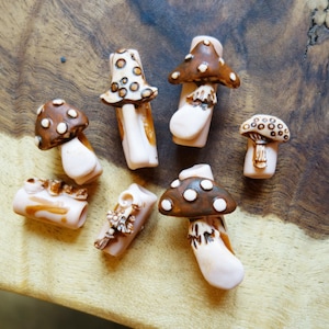 May include: A set of six ceramic mushroom-shaped charms in shades of brown and white with white polka dots. The charms are designed to look like mushrooms with a stem and a cap.