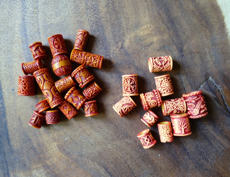 May include: A collection of wooden hair beads with intricate carvings and a reddish-brown color. Some beads have a lighter brown color with a more intricate design.