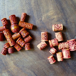 May include: A collection of wooden hair beads with intricate carvings and a reddish-brown color. Some beads have a lighter brown color with a more intricate design.