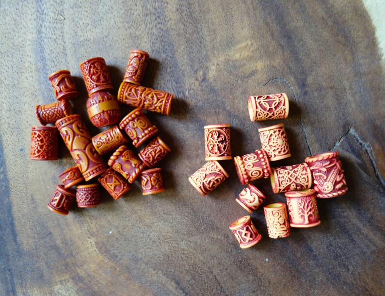 May include: A collection of wooden hair beads with intricate carvings and a reddish-brown color. Some beads have a lighter brown color with a more intricate design.