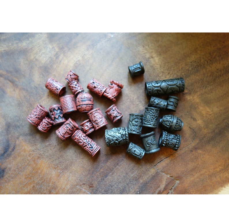 May include: A collection of hair beads in two colors: dark red and black. The beads are cylindrical and have intricate designs carved into their surfaces.