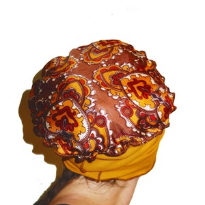 May include: A mustard yellow turban with a paisley patterned top in shades of brown, orange, and white. The turban has a ruffled edge and is made of soft fabric. The head covering is a stylish accessory.