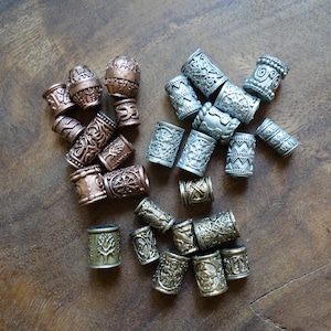 May include: A collection of silver, copper, and bronze hair beads with intricate designs. The beads are arranged on a wooden surface.