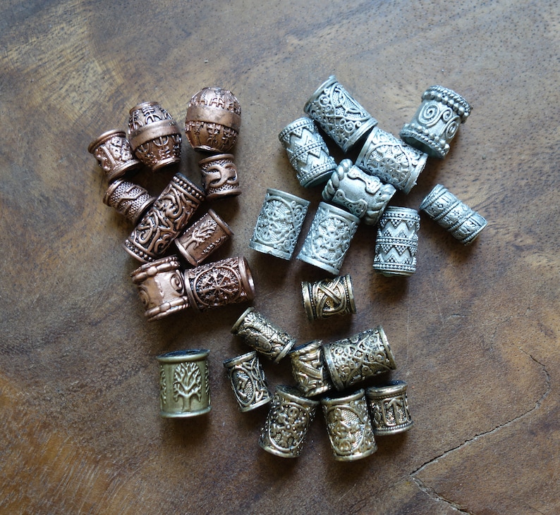 May include: A collection of silver, copper, and bronze hair beads with intricate designs. The beads are arranged on a wooden surface.