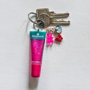 Puede incluir: Un llavero con llaves, un tubo de brillo labial brillante Essence Juicy Bomb y abalorios. El brillo labial es rosa con el nombre de la marca y del producto impreso. Los abalorios incluyen un osito de goma rosa, una mariposa roja y una concha azul.