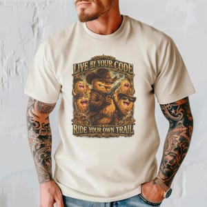 May include: Beige t-shirt featuring a graphic of a peanut cowboy holding a gun, wearing a hat and jacket. The text "LIVE BY YOUR CODE" and "RIDE YOUR OWN TRAIL" are above and below the graphic.