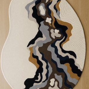 Könnte beinhalten: Ein cremefarbener, ovaler Teppich mit einem einzigartigen Design. Der Teppich zeigt ein Muster aus fließenden Formen in Schwarz-, Braun-, Grau- und Goldtönen. Der Text "AMAN RUGS" ist in den Ecken oben links und unten links zu sehen.