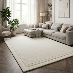 May include: A large, rectangular, off-white area rug featuring a subtle, double-lined border design. The rug has a soft, plush texture and is positioned on a dark brown herringbone wood floor in a living room setting with a sofa and plants.