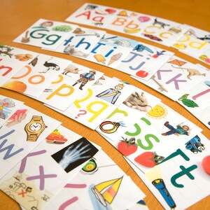 Puede incluir: Un conjunto de tarjetas de aprendizaje del alfabeto con ilustraciones y letras coloridas. Cada tarjeta presenta una letra diferente del alfabeto y una imagen correspondiente. Las tarjetas est&aacute;n dispuestas en semic&iacute;rculo sobre una superficie de madera.