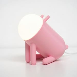 Könnte beinhalten: Eine rosa Novelty-Lampe mit zylindrischem Körper und vier beinartigen Stützen. Die Lampe hat oben ein rundes, leuchtendes weißes Licht und ein kleines schwanzartiges Detail auf der Rückseite. Ein weißes Netzkabel erstreckt sich von der Basis.