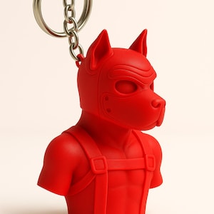 Op de afbeelding: Een rode sleutelhanger met een gestileerde hondenbuste, met een masker en harnas. De sleutelhanger heeft een zilveren ketting en ring voor bevestiging. De hond heeft spitse oren en een prominente snuit.
