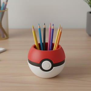 Peut inclure: Un porte-crayons en forme de Pokéball rouge, blanc et noir rempli de crayons de couleur. Les crayons sont jaunes, bleus, noirs, roses et oranges. Le porte-crayons est posé sur une surface en bois clair.