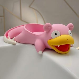 May include: A pink Slowpoke-shaped soap dish with a yellow beak and red mouth. The dish has a curved tail and small ears. The product is designed to sit on the edge of a sink or tub.