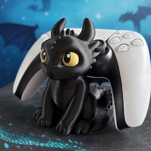 Könnte beinhalten: Ein schwarzer Controller-Halter im Drachen-Design mit gelben Augen und einem weiß-schwarzen Controller. Der Drache sitzt vor dem Controller. Der Hintergrund ist ein blauer Farbverlauf mit Drachensilhouetten.