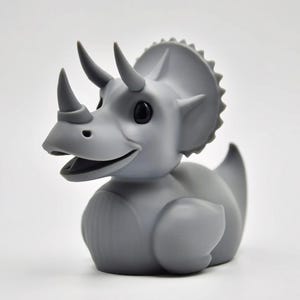 Pode incluir: Um pato de borracha cinzento em forma de triceratops com olhos pretos, três chifres e uma gola. O pato dinossauro tem um bico aberto e foi concebido para flutuar na água. Um brinquedo de banho divertido.