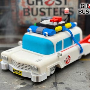 Può includere: Un'auto giocattolo bianca con finestrini blu e una pinna rossa e bianca, che ricorda l'Ecto-1 di Ghostbusters. L'auto ha un portapacchi nero con dettagli rossi e gialli, e il logo Ghostbusters è visibile sul lato. La parte anteriore dell'auto recita "ECTO-1".