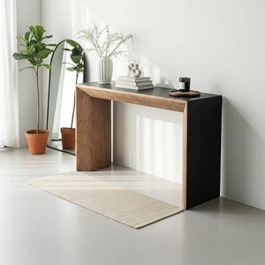 Op de afbeelding: Een moderne console met een zwart blad en een houten frame. Een witte vaas met bloemen, boeken en een kaars staan op de tafel. Een beige tapijt ligt op de vloer en een plant in pot en een spiegel staan op de achtergrond.