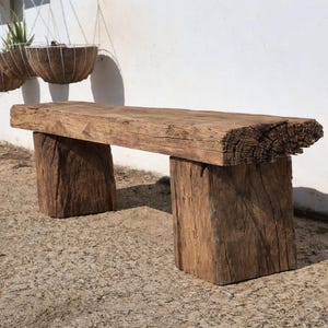 May include: A weathered wooden bench with a natural, rustic aesthetic. The bench has a thick, rectangular seat supported by two square legs. The wood displays a warm brown hue, highlighting the natural grain and texture. The bench is approximately 150 cm long.