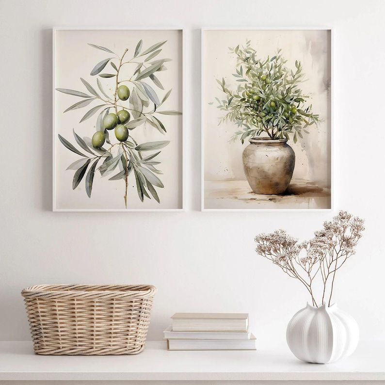 May include: Two framed watercolor prints. One features an olive branch with green olives and gray-green leaves. The other depicts a vase filled with green foliage. A woven basket, books, and a white vase with dried flowers are on a white surface.