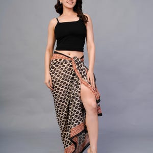 Pode incluir: Um top de camisola preto combinado com um sarongue estampado. O sarongue apresenta um padrão de folhas pretas e bege repetidas com uma borda vermelha. A modelo está descalça, com o sarongue amarrado na cintura e uma fenda alta.