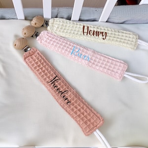 以下が含まれることがあります： クリーム、ピンク、ピーチ色のワッフル生地の、パーソナライズされたベビーおしゃぶりクリップ3つ。各クリップには木製のビーズと、対応する色で刺繍された名前「Henry」、「River」、「Theodore」が付いています。クリップは白い表面に置かれています。