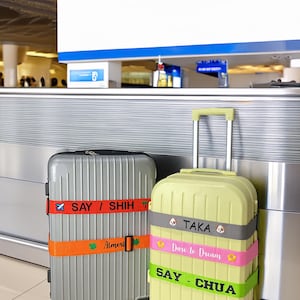 Könnte beinhalten: Zwei Koffer an einem Flughafen. Einer ist silberfarben mit einem orangefarbenen Riemen mit der Aufschrift "SAY / SHIH" und "Almeri". Der andere ist gelb mit Riemen mit der Aufschrift "TAKA", "Dare to Dream" und "SAY - CHUA".