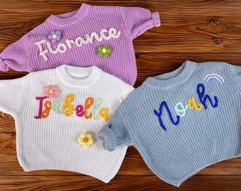 Op de afbeelding: Drie gebreide truien in lavendel, wit en blauw, elk met een naam op de voorkant geborduurd. De lavendelkleurige trui zegt "Florance", de witte trui zegt "Isabella" en de blauwe trui zegt "Noah". Elke trui heeft bloemversieringen.