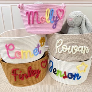 Peut inclure: Cinq paniers de rangement en corde tressée de différentes couleurs, chacun avec un nom brodé sur le devant. Les noms incluent "Molly", "Comet", "Rowan", "Finley" et "Jenson". Un lapin en peluche gris est dans l'un des paniers.