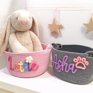 May include: Two handmade rope baskets, one pink and one gray, sit side-by-side. The pink basket has the name "Lottie" in colorful letters and a flower. The gray basket has the name "Asha" and a paw print. A stuffed bunny is in the pink basket.