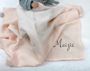 Puede incluir: Una manta de bebé de tejido de gofre en tonos rosa claro y blanco. La manta está drapeada, con el nombre "Maizie" escrito en negro. La manta está hecha de un material texturizado.