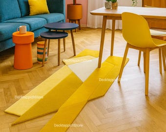 Può includere: Un soggiorno moderno con un divano blu, accenti gialli e un tappeto geometrico giallo. La stanza presenta un tavolo e una sedia in legno, una grande pianta in vaso e una finestra.