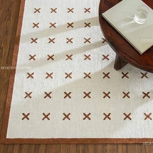 Puede incluir: Una alfombra rectangular con base blanca, un patrón repetitivo de pequeños símbolos de cruz en color marrón y un borde marrón sólido. La alfombra está sobre un suelo de madera, parcialmente cubierta por una mesa redonda de madera oscura.