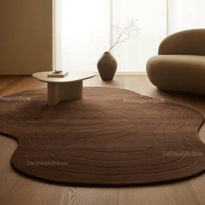 May include: An irregular, organic-shaped area rug in a deep chocolate brown color, featuring a series of concentric, wavy, carved-in lines that mimic topographical patterns. The rug lies flat on a light-toned wooden floor surface.
