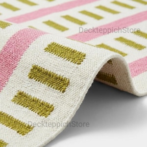 May include: A rolled rug with a geometric pattern. The rug has alternating stripes of pink and white, with rectangular olive green shapes. The rug is made of a textured material, and the design is visible on the rolled edge.