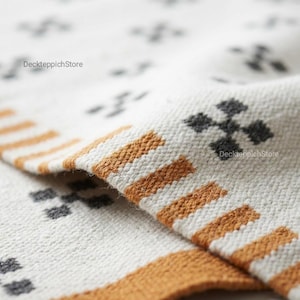 Könnte beinhalten: Ein gewebtes Textil mit cremeweißer Basis und einem sich wiederholenden Muster aus kleinen, dunkelgrauen kreuzartigen Formen. Der Rand zeigt eine Bordüre aus dicken, horizontalen senfgelben Streifen. Der Text DeckteppichStore ist sichtbar.